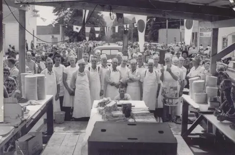 A historic photograph of a group of men in aprons standing behind tables with large stacks of plates.