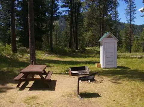 A picnic table, grill, and outhouse in a wooded area.