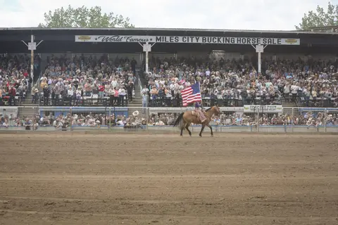 A person rides a horse carrying an American flag in an arena before a large crowd.