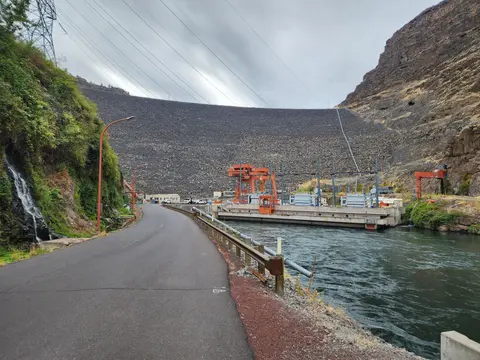 A paved road runs along a river next to a large rock dam.
