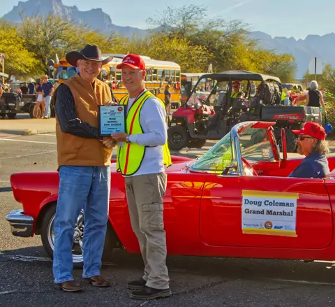 Two men stand next to a red convertible holding a plaque near vehicles and mountains.