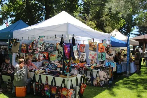 A vendor booth at an outdoor market displays many colorful bags and totes under a white canopy.