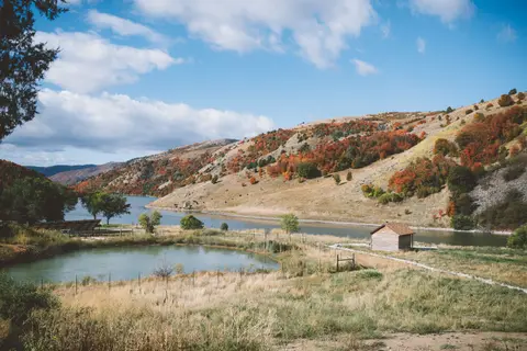 A scenic landscape view featuring a lake, hills with autumn-colored trees, and a small wooden cabin.