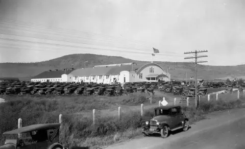 Historic black and white exterior view of a large building surrounded by parked vintage cars.