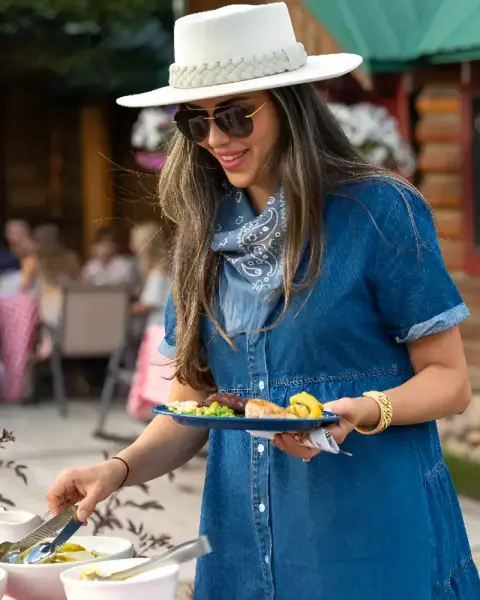 A woman in a white hat and denim dress serves herself food from a buffet.