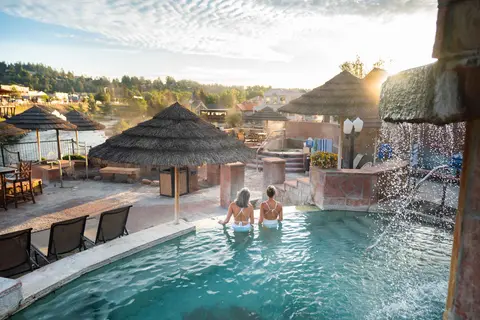 Two women relax in an outdoor hot spring pool with thatched umbrellas nearby.