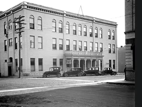 A historic photograph showing a large building exterior with vintage automobiles parked in front.