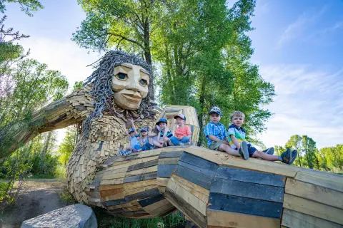 Several children sit on a large wooden troll sculpture outdoors.