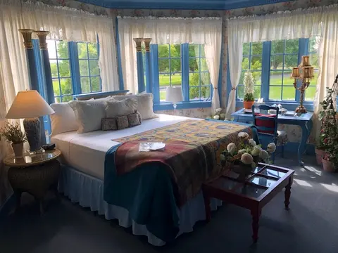 A brightly lit bedroom with a large bed, blue window frames, and sheer curtains.