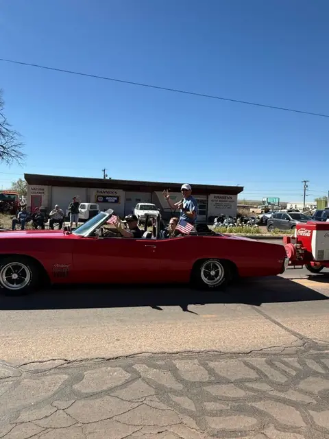 People ride in a red convertible car during an outdoor event.