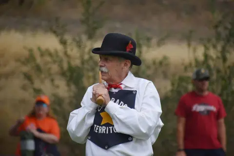 A man wearing a bowler hat and historical attire holds a wooden bat.