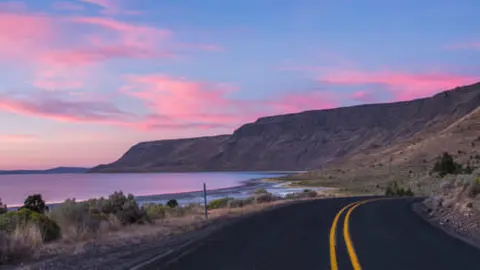 A winding road runs alongside a lake and hills beneath a pink sunset sky.
