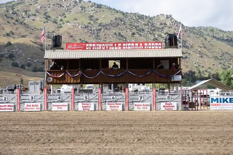 A rodeo arena with a wooden announcer stand and numbered chutes against a mountainous backdrop.