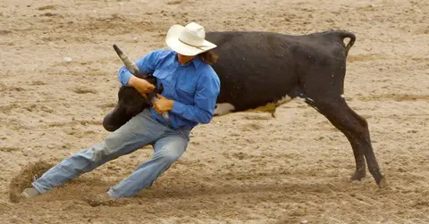A cowboy wrestles a steer in a dirt arena.