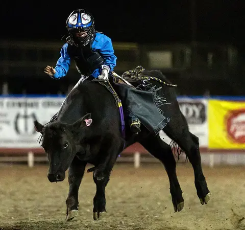A young rider wearing a helmet on a bucking bull in an arena.