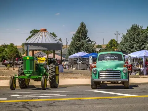 A vintage tractor and a classic truck parked near a street market with tents.