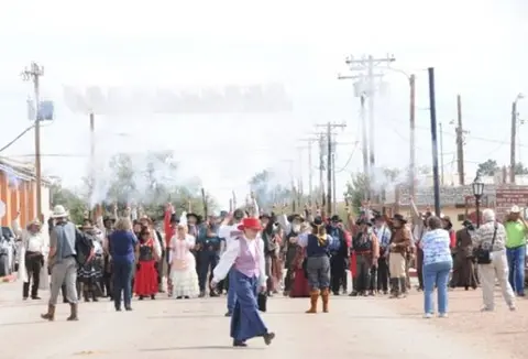 A crowd of people in western attire walking down a dusty street with some smoke.