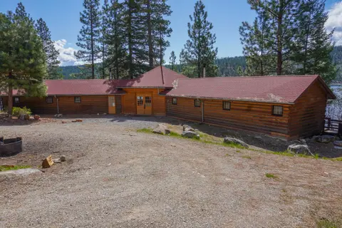 A log building with a red shingled roof stands amidst trees with a dirt area in front.