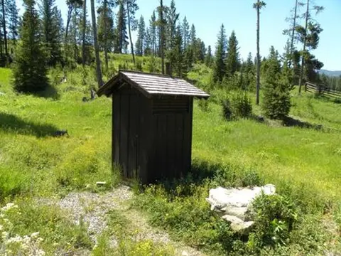 A small wooden outhouse stands in a grassy clearing surrounded by trees.