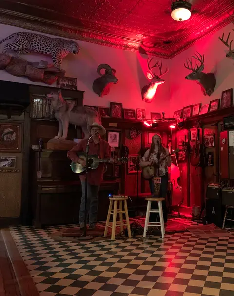 Two musicians perform on a stage in a rustic room decorated with taxidermy and checkered floors.