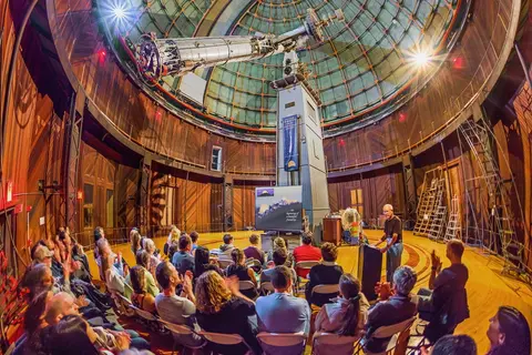 A presentation is being given inside a large domed observatory with a telescope.