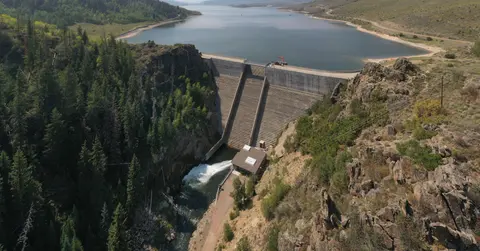 An aerial view of a dam and reservoir surrounded by forested mountains and rocky terrain.