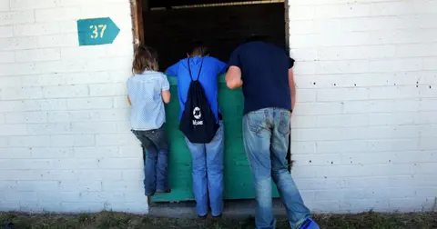 Three people look over a green half-door into a white brick stable.