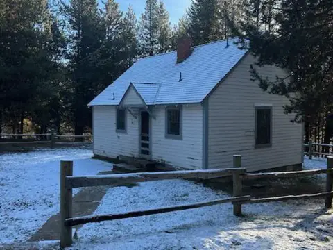 A small white house sits surrounded by trees with snow on the ground.