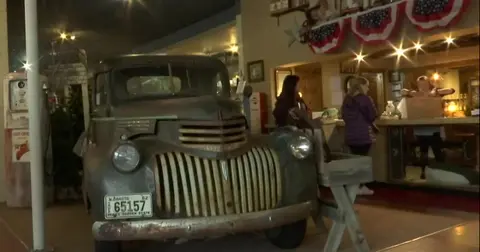 A vintage truck is parked inside a rustic building with people standing in the background.