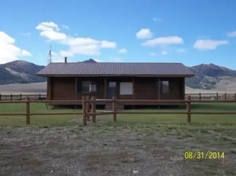 A wooden cabin stands in a fenced yard with mountains in the background.