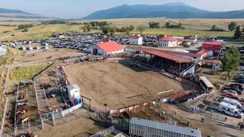 An aerial view shows a rodeo arena with livestock pens, stands filled with spectators, and surrounding fairgrounds.