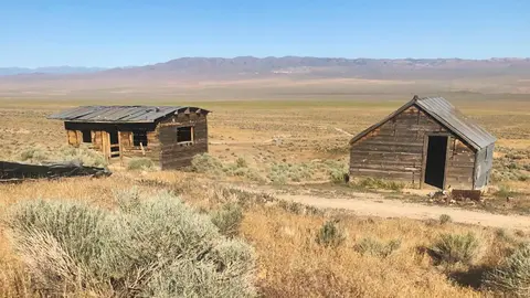 Two weathered wooden cabins stand in a dry desert landscape with distant mountains.