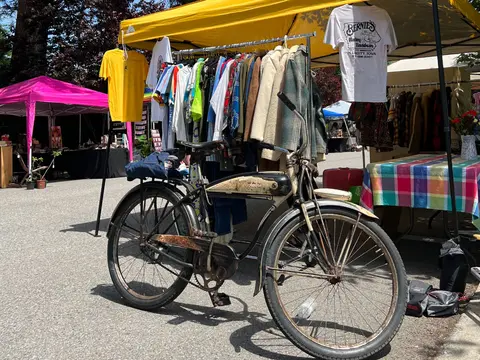 An old bicycle stands parked in front of a clothing rack at an outdoor market.