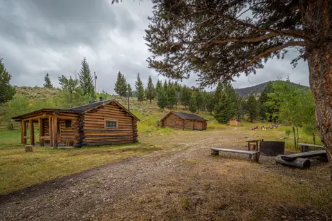Log cabins sit in a clearing surrounded by trees and mountains under a cloudy sky.
