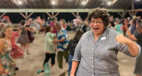 A woman celebrating joyfully while people dance in the background inside a hall.