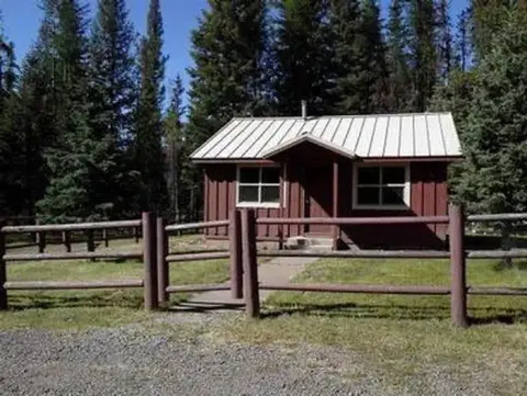 A small cabin stands surrounded by pine trees and a wooden fence.