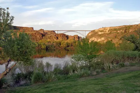 A bridge spans across a canyon over a river with trees and grass in the foreground.