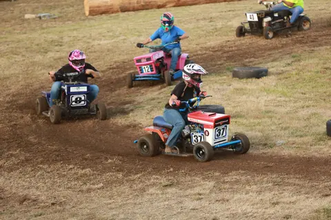 People race modified lawn mowers on a dirt track.