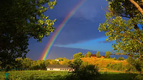 A vibrant rainbow arches across a dark blue sky over a rural landscape with buildings and trees.
