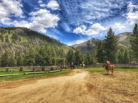 People ride horses along a dirt path near a wooden fence with mountains in the background.