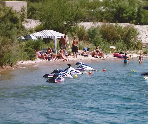 People relaxing on a sandy beach by a body of water with watercraft moored nearby.
