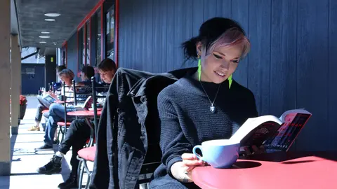 A woman reads a book while sitting at an outdoor table with a cup.