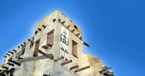 An exterior view of a stucco building with exposed wooden beams and a painted figure banner against a clear blue sky.