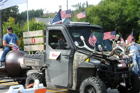 A decorated utility vehicle towing a trailer with a man sitting on it during a parade.