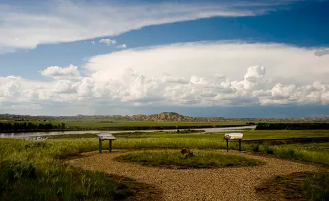 A wide view of a grassy plain and river under a dramatic cloudy sky.