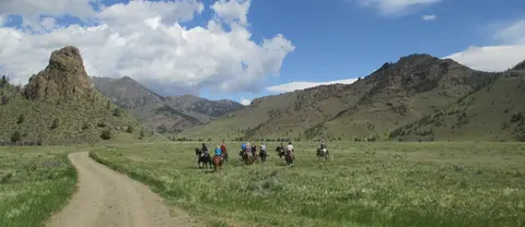 A group of people ride horses through a green valley with mountains in the background.
