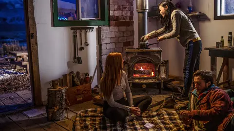 Three people inside a cabin, with one person cooking on a wood stove.
