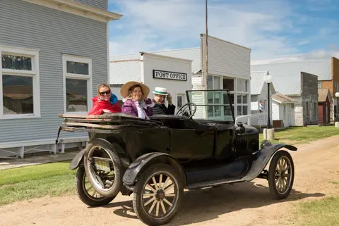 Three people in period attire sit in an antique car on a historic town street.