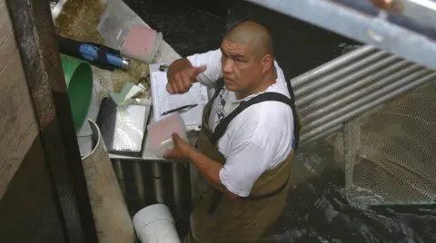 A man working near a water structure, holding a small container and pointing upward.