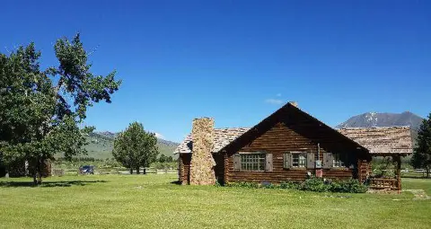 A rustic log cabin sits on a grassy lawn with mountains and trees in the background.